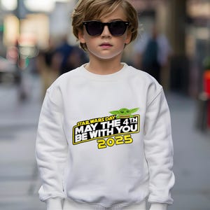 May include: White sweatshirt featuring a Star Wars Day graphic with the text "May the 4th Be With You 2025" in yellow and black. The design includes a green Grogu character. The sweatshirt is a casual, long-sleeved garment.
