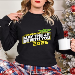 May include: Black sweatshirt featuring a graphic of Baby Yoda and the text "STAR WARS DAY MAY THE 4TH BE WITH YOU 2025" in yellow and white. The sweatshirt is paired with plaid pajama pants. A white mug is held in the hand.