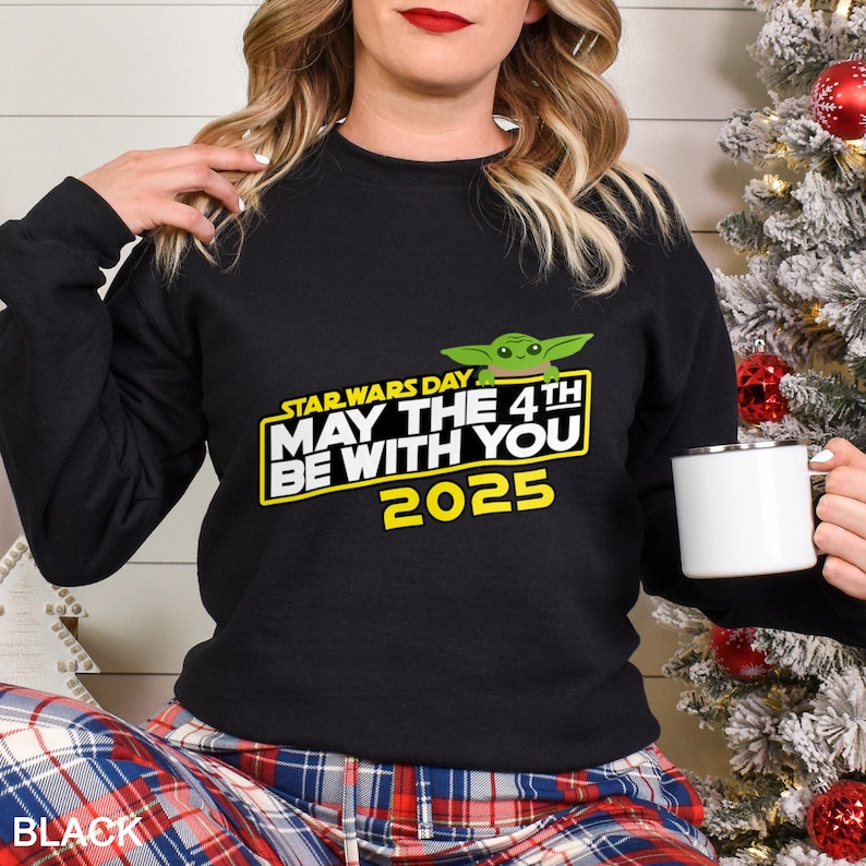 May include: Black sweatshirt featuring a graphic of Baby Yoda and the text "STAR WARS DAY MAY THE 4TH BE WITH YOU 2025" in yellow and white. The sweatshirt is paired with plaid pajama pants. A white mug is held in the hand.