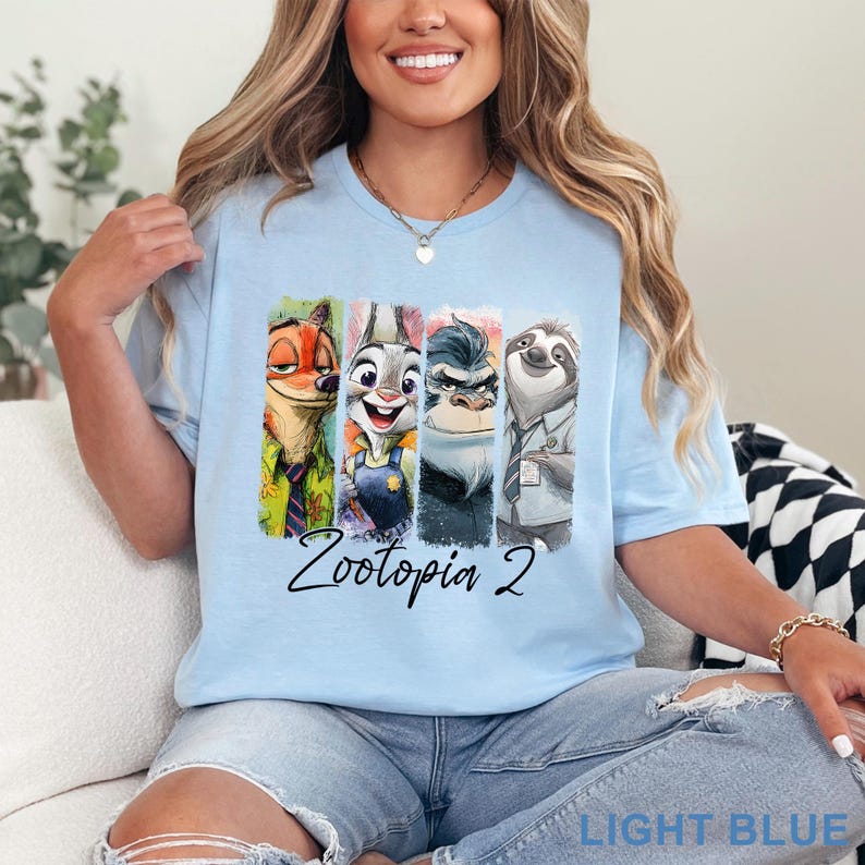 Peut inclure: T-shirt bleu clair avec les personnages de Zootopie. Le design pr&eacute;sente Nick Wilde, Judy Hopps, le chef Bogo et Flash dans des panneaux verticaux color&eacute;s, avec "Zootopia 2" &eacute;crit en dessous. Les mots "LIGHT BLUE" sont en bas.