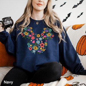 Puede incluir: Sudadera azul marino con un dise&ntilde;o floral de Mickey Mouse. El arreglo floral incluye una variedad de flores coloridas. La palabra "Navy" est&aacute; impresa en la parte inferior de la imagen. La sudadera es parte de una colecci&oacute;n tem&aacute;tica de oto&ntilde;o o Halloween.