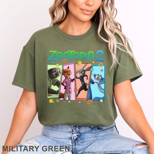 May include: Military green t-shirt with the text "ZOOTOPIA 2" and a graphic of characters from the animated film. The graphic features a sloth, fox, rabbit, and snake in colourful rectangular frames. The shirt is a solid colour.
