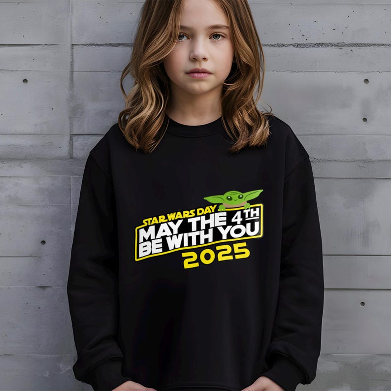 May include: Black sweatshirt featuring the text "STAR WARS DAY MAY THE 4TH BE WITH YOU 2025" in yellow and white, with a green Grogu graphic. The sweatshirt is worn by a person with long, wavy brown hair.