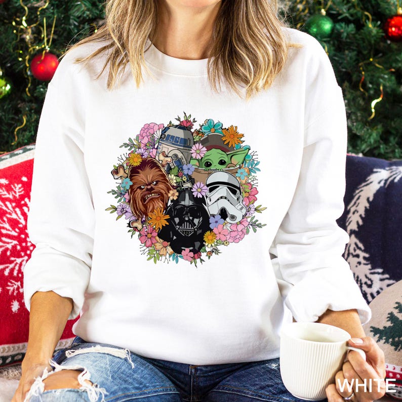 Puede incluir: Sudadera blanca con un dise&ntilde;o floral colorido que presenta personajes de Star Wars: Darth Vader, Chewbacca, R2-D2, un Stormtrooper y Grogu. El dise&ntilde;o est&aacute; rodeado de flores en tonos rosas, naranjas y azules.