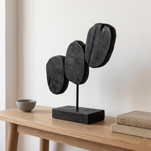 May include: A modern black wooden sculpture with three oval shapes mounted on a black metal stand and a rectangular base. The sculpture is displayed on a light wood table, with a small gray bowl and two books nearby.
