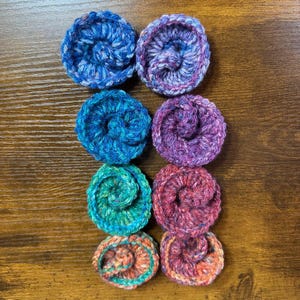 May include: Eight handmade crochet coasters in various colors, including blue, purple, green, and red. Each coaster features a spiral design and is made of textured yarn. The coasters are arranged on a wooden surface.