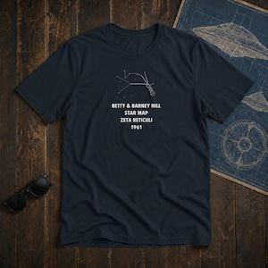 Pode incluir: Uma t-shirt azul-marinho de manga curta com um gráfico branco de um mapa estelar e o texto: BETTY & BARNEY HILL STAR MAP ZETA RETICULI 1961. A peça possui gola redonda e um corte padrão, exibindo um design de constelações celestiais.