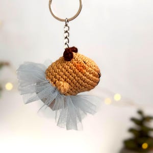 May include: A crocheted capybara wearing a light blue tulle tutu, attached to a silver key chain.