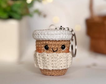 May include: A crocheted coffee cup keychain with a smiling face and a white crocheted cozy. The keychain is attached to a silver key ring.