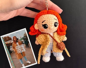 May include: A crocheted keychain featuring a girl with red hair, wearing a yellow jacket and blue jeans. The keychain is attached to a silver key ring.