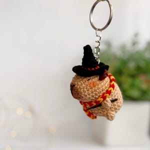 May include: A crocheted capybara wearing a black witch hat and a red and yellow scarf, holding a wand. The capybara is attached to a silver key chain.