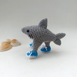 May include: A crocheted gray shark toy with black eyes and white accents. The shark is wearing blue sneakers with white laces. Seashells are in the background. The toy is handmade.