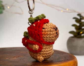 May include: A crocheted stuffed animal keychain shaped like a dog wearing a red and green strawberry costume. The keychain is attached to a silver key ring.