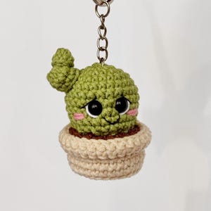 May include: A green crocheted cactus with a smiling face and large black eyes, wearing a pink blush. The cactus is in a beige crocheted pot and attached to a silver keychain.