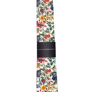 Pode incluir: Uma gravata floral com fundo branco e um padr&atilde;o colorido de flores vermelhas, amarelas, azuis e verdes. A gravata &eacute; embrulhada com uma faixa preta que diz "POMP & CEREMONY".