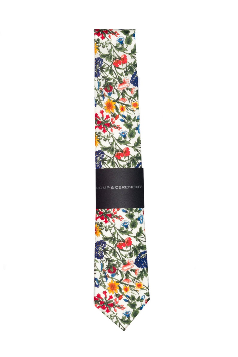 Pode incluir: Uma gravata floral com fundo branco e um padr&atilde;o colorido de flores vermelhas, amarelas, azuis e verdes. A gravata &eacute; embrulhada com uma faixa preta que diz "POMP & CEREMONY".