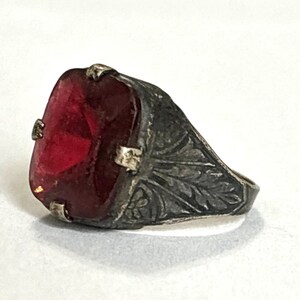 May include: Antique silver ring featuring a large, rectangular, deep red gemstone. The ring's band is intricately detailed with a leaf pattern, adding a vintage aesthetic. The gemstone is securely set within the ring's frame.
