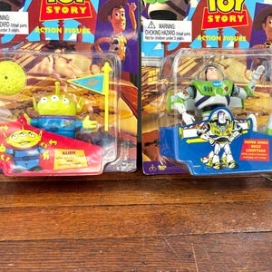 Może przedstawiać: Dwie figurki akcji Toy Story w oryginalnym opakowaniu. Jedna przedstawia postać Obcego z ż&oacute;łtym księżycem i flagą. Druga to Buzz Lightyear. Opakowanie zawiera tekst "ACTION FIGURE" i ostrzeżenie o niebezpieczeństwie uduszenia.
