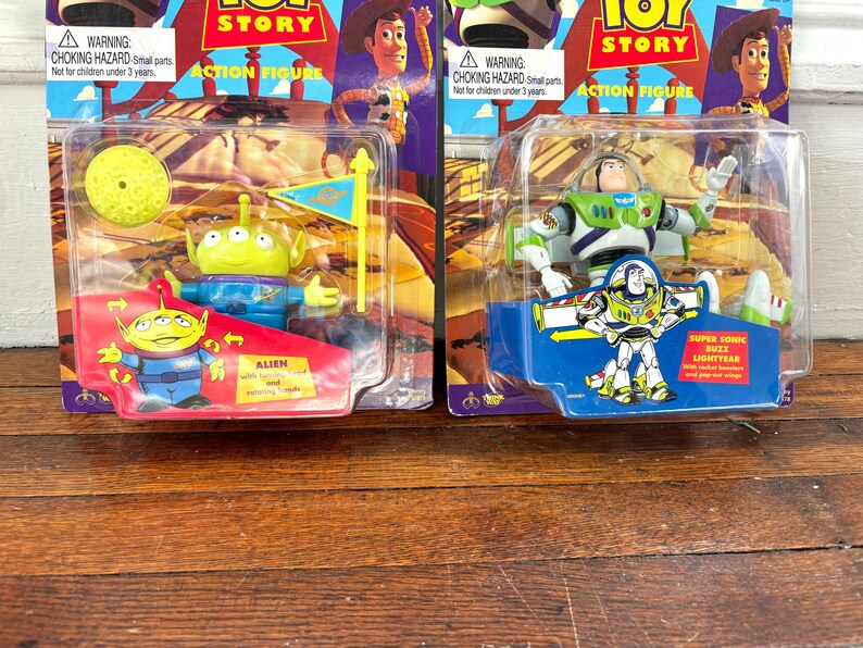 Może przedstawiać: Dwie figurki akcji Toy Story w oryginalnym opakowaniu. Jedna przedstawia postać Obcego z ż&oacute;łtym księżycem i flagą. Druga to Buzz Lightyear. Opakowanie zawiera tekst "ACTION FIGURE" i ostrzeżenie o niebezpieczeństwie uduszenia.