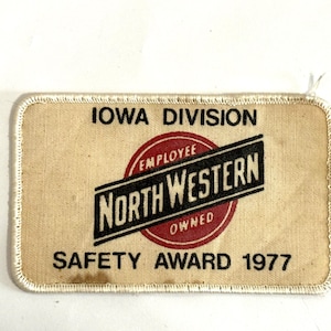 May include: Vintage rectangular patch with the text "IOWA DIVISION" at the top and "SAFETY AWARD 1977" at the bottom. The center features a red circle with "EMPLOYEE OWNED" and "NORTH WESTERN" in a black and white design.