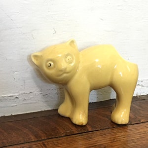May include: A ceramic cat figurine in a light yellow color. The cat is standing with its head turned slightly to the left. The figurine has a glossy finish and is placed on a wooden surface.