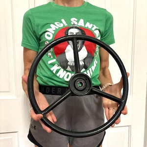 May include: A black, circular, three-spoke steering wheel with a central hub featuring a hollow, circular opening. The steering wheel has a smooth, solid rim and three thin, straight spokes that connect the outer ring to the center hub.