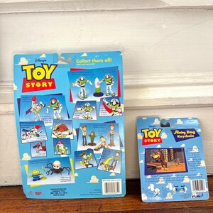 Peut inclure: Deux porte-cl&eacute;s Toy Story sur un emballage bleu. L'un repr&eacute;sente Buzz l'&Eacute;clair et l'autre Slinky Dog. L'emballage comprend le logo Toy Story et des illustrations de personnages. Les porte-cl&eacute;s sont pr&eacute;sent&eacute;s sur une surface en bois.
