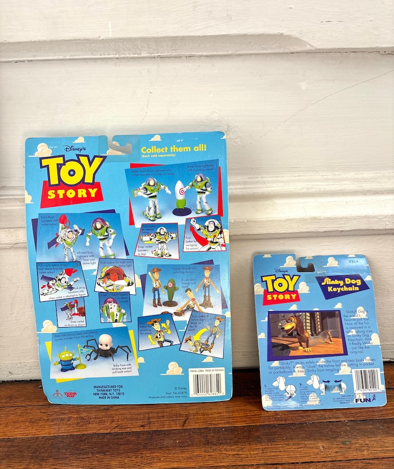 Peut inclure: Deux porte-cl&eacute;s Toy Story sur un emballage bleu. L'un repr&eacute;sente Buzz l'&Eacute;clair et l'autre Slinky Dog. L'emballage comprend le logo Toy Story et des illustrations de personnages. Les porte-cl&eacute;s sont pr&eacute;sent&eacute;s sur une surface en bois.