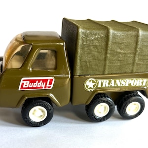 May include: Olive green toy truck with a canvas-covered bed. The truck has black tires with white rims and the text "Buddy L" in a red and white box. The side of the truck bed reads "TRANSPORT" in white.