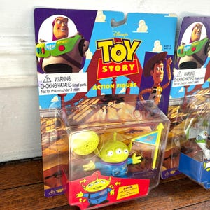 Może przedstawiać: Zapakowane figurki akcji Toy Story z Buzzem Astralem, Chudym i Obcym. Figurka Obcego jest w przezroczystym plastikowym etui z ż&oacute;łtym księżycem i flagą. Opakowanie zawiera tekst "Toy Story Action Figure".