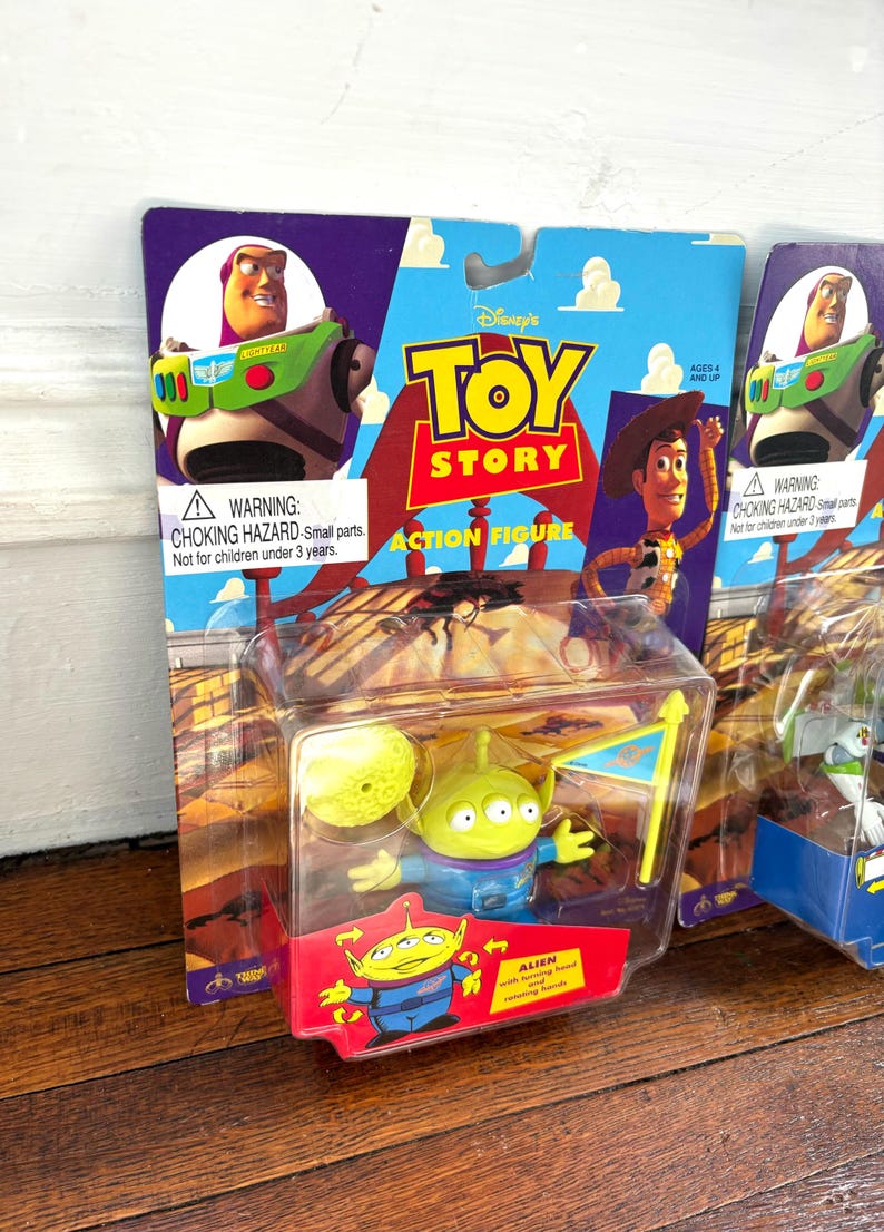 Może przedstawiać: Zapakowane figurki akcji Toy Story z Buzzem Astralem, Chudym i Obcym. Figurka Obcego jest w przezroczystym plastikowym etui z ż&oacute;łtym księżycem i flagą. Opakowanie zawiera tekst "Toy Story Action Figure".
