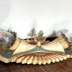 May include: A dual-light wall sconce featuring a cream-colored base with a fan-shaped design, teal and rust-colored accents, and a central decorative finial. Two iridescent, flame-shaped light bulbs are set into fluted, trumpet-like holders.