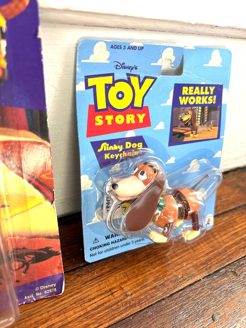 Peut inclure: Un porte-cl&eacute;s Slinky Dog de Toy Story dans son emballage d'origine. L'emballage est bleu avec le logo Toy Story et l'inscription &laquo; REALLY WORKS! &raquo;. Le Slinky Dog est marron et beige. L'emballage indique &laquo; AGES 5 AND UP &raquo;.