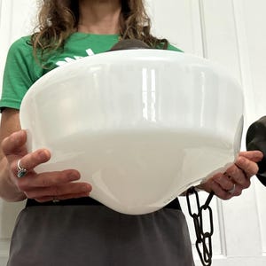 May include: A large, white, bowl-shaped glass light fixture shade with a smooth, opaque finish. The shade features a dark, metallic brown circular mounting base on one side and a short, dark, metallic brown chain link attached to the bottom.