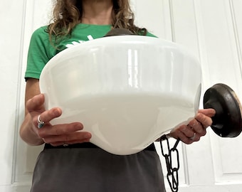 May include: A large, white, bowl-shaped glass light fixture shade with a smooth, opaque finish. The shade features a dark, metallic brown circular mounting base on one side and a short, dark, metallic brown chain link attached to the bottom.