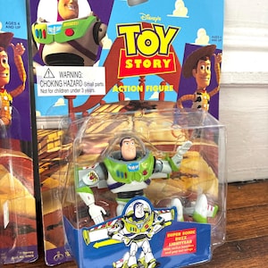 Może przedstawiać: Figurka Buzz Lightyear z filmu Toy Story Disneya, zapakowana na niebiesko-fioletowej karcie. Figurka znajduje się w przezroczystym plastikowym etui. Karta przedstawia Woody'ego i Buzza Lightyeara. Opakowanie zawiera ostrzeżenie o małych częściach.