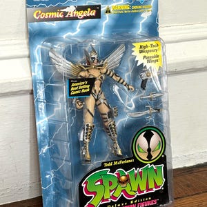 May include: A plastic package containing a Cosmic Angela action figure from the Spawn Deluxe Edition Ultra-Action Figures line. The figure is a female character with white wings, a gold and black costume, and a gun. The package also includes a small comic book.