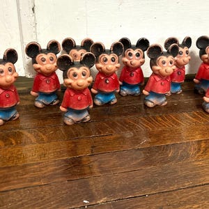 May include: A collection of vintage Mickey Mouse figurines. Each figurine is wearing a red shirt, blue shorts, and black ears. The figurines are standing on a wooden surface.