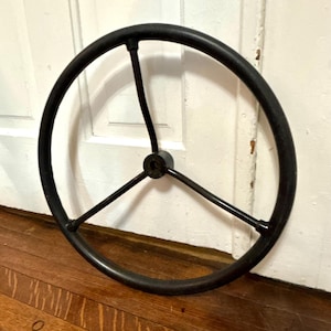 May include: A circular black steering wheel featuring a three-spoke design that connects to a central hub. The wheel has a smooth, matte finish and a consistent thickness throughout the outer rim and the three metal spokes extending to the center.
