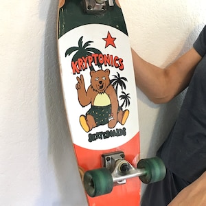 May include: A vintage Kryptonics skateboard with a bear graphic giving a peace sign. The board has orange and green wheels, a red star, and the words "Kryptonics Skateboards". The board is a retro style.