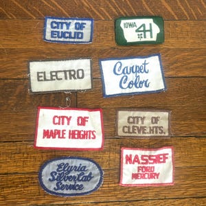 May include: A collection of embroidered patches with various text. The patches are rectangular and oval, with text such as "CITY OF EUCLID", "ELECTRO", "Carpet Color", "CITY OF MAPLE HEIGHTS", "Elyria Silver Cab Service", and "NASSIEF FORD MERCURY".