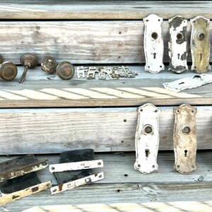 May include: A collection of vintage door hardware including several round brass knobs, rectangular metal backplates with keyholes in white and distressed finishes, and black metal door latches arranged on a wooden surface.