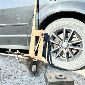 May include: A heavy-duty, rusted iron post vise featuring a long vertical mounting bar, a screw-operated clamping mechanism with a spherical handle, and a separate rectangular wooden base block with a circular metal mounting plate attached to the top.