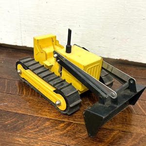 May include: A yellow toy bulldozer featuring black rubber tracks, a black front blade, and a black exhaust stack. The construction vehicle has a yellow metal body with a cab area and silver-toned metal hardware connecting the blade to the main frame.