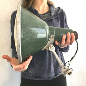 May include: Vintage green metal spotlight with a white rim. The spotlight has a long handle and a bracket for mounting. The spotlight is held by a person. The spotlight has a vintage, industrial aesthetic.