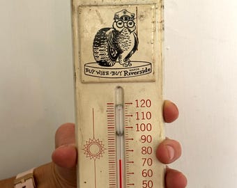 May include: A vintage off-white metal thermometer featuring a black and white graphic of an owl wearing a hat and glasses, leaning on a tire. The text reads Buy Wise-Buy Riverside Wards. It displays Fahrenheit temperatures from -10 to 120.