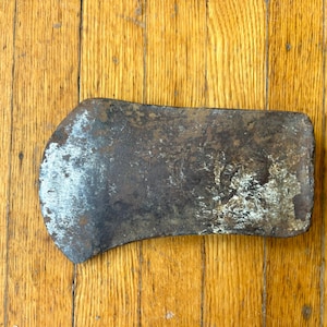 May include: A weathered metal axe head featuring a dark, oxidized surface with patches of rust and a worn, metallic edge. The tool displays signs of heavy use and age, with a curved blade profile and a rectangular poll, showing a textured finish.