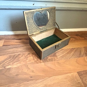 May include: An open, antique jewelry box with a heart-shaped mirror on the inside of the lid. The box is rectangular with a brass finish and a green velvet lining. The lid is open, revealing the interior.