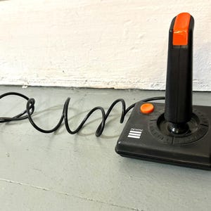 May include: A black video game joystick with a square base and a vertical handle. The handle features a bright orange button at the top, and the base includes a smaller orange button and the word GEMINI printed in white with three horizontal lines.
