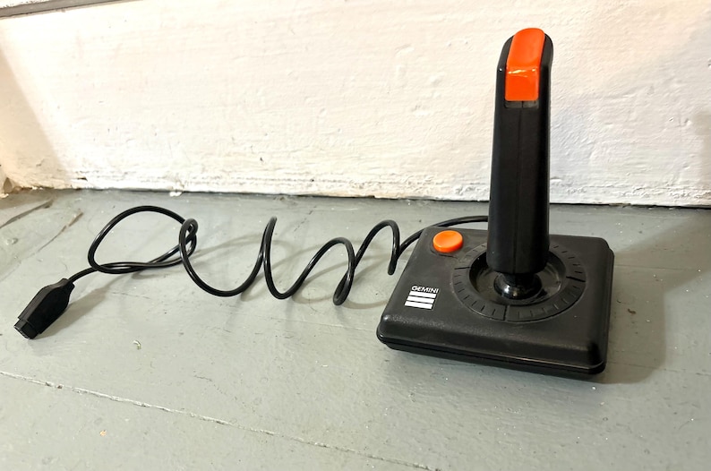May include: A black video game joystick with a square base and a vertical handle. The handle features a bright orange button at the top, and the base includes a smaller orange button and the word GEMINI printed in white with three horizontal lines.